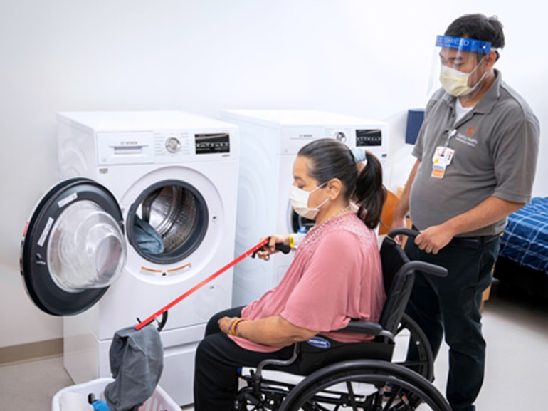 A woman in a wheelchair using a trigger grabber to pull laundry from a basket and into a washing machine.