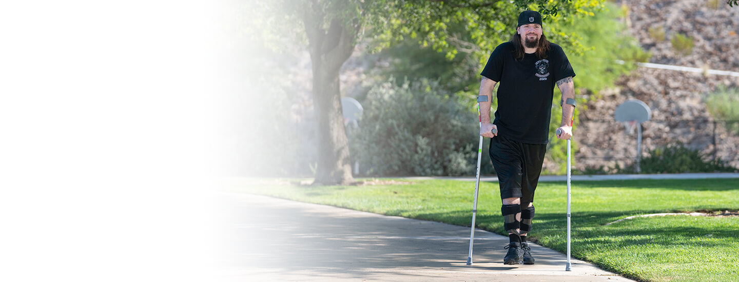A young man wearing a black shirt and ballcap using forearm crutches to walk outside on a sidewalk.