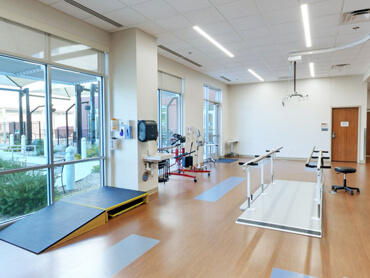 The interior of a physical therapy gym with equipment facing windows looking out the a courtyard.