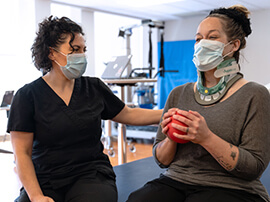 Woman wearing a face mask and a neck brace and squeezing a small red therapy ball.