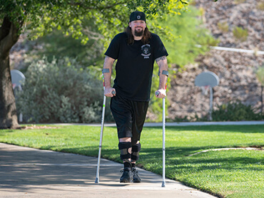 Man dressed in all black wearing braces on his calves and using forearm crutches to walk in a park on a sunny spring day.