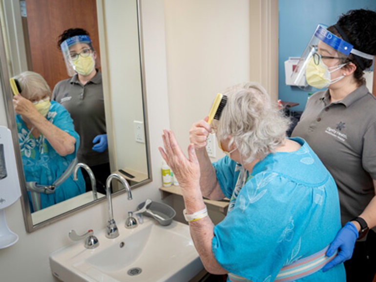 Female therapist standing next to an older woman brushing her white hair and looking into a mirror.