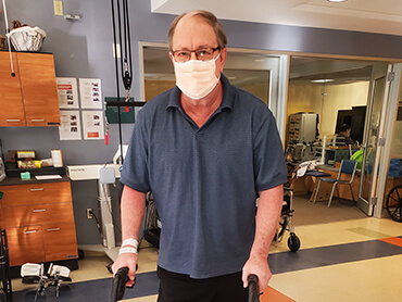 Man wearing glasses and a face mask in a therapy room using a walker for support.