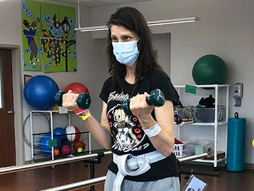 A woman with long, dark hair doing bicep curls with small hand weights in a therapy gym.