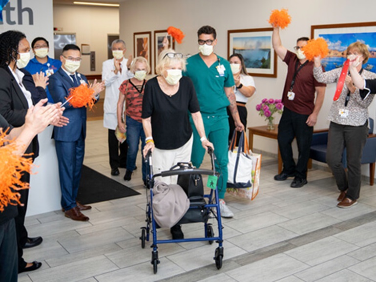 woman using rolling walker leaving hospital with hospital staff cheering her on.