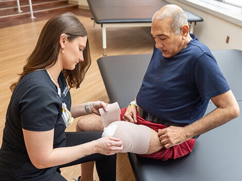 A therapist assists a patient to wrap his leg amputation in the therapy gym.