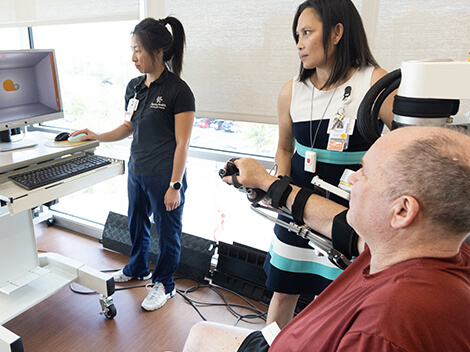 A male patient sits in a wheelchair and with the help of his care team utilize technology to regain arm motion and accuracy.