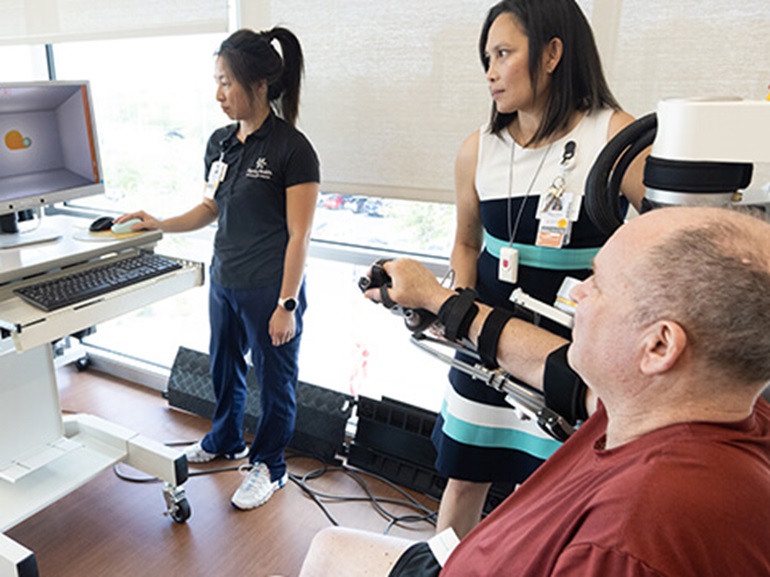 A male patient sits in a wheelchair and with the help of his care team utilize technology to regain arm motion and accuracy.