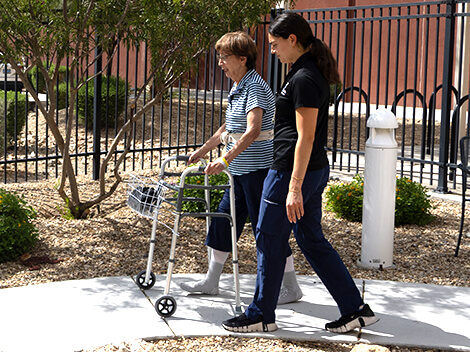 A women walks using a walker down the side walk with her therapist.