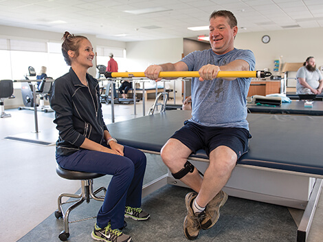 A patient smiles while working on arm exercises in the therapy gym with his therapist beside him.
